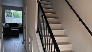 Un escalier moderne avec une rampe noire, des marches en bois clair et une vue sur un salon avec un canapé de couleur foncée et une fenêtre équipée de stores.