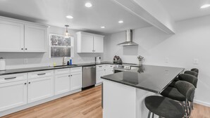 A modern kitchen with white cabinetry, a central island with bar stools, stainless steel appliances, and a large window with a decorative metal grid. - Tennessee Tranquility • Fire Pit • Forest Views (Ashland City)