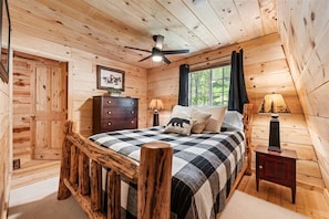 Een slaapkamer in een houten chalet, met een bed, een dressoir, nachtkastjes, een plafondventilator en een raam met gordijnen.