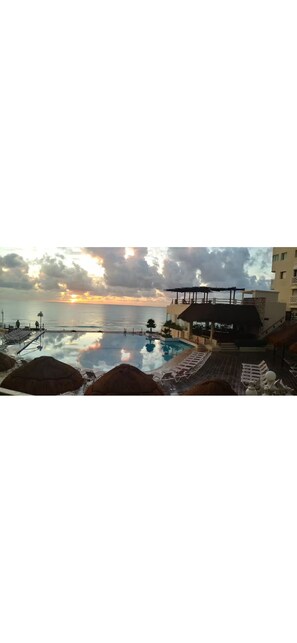 Una piscina con sillones de descanso y sombrillas, un atardecer al fondo y edificios a lo lejos.