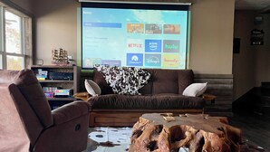 Una sala de estar con un sofá marrón, un sillón marrón y una mesa de centro de madera. Una alfombra de piel de vaca. Una pantalla de proyector muestra la interfaz de Roku con varias opciones de streaming.