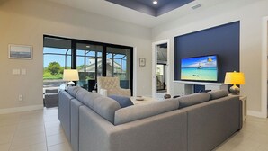A modern living room with a grey sectional sofa, a flat-screen TV mounted on the wall, and a large window offering a view of the outdoors. - Cape Luxe Canal Retreat - Saltwater Pool & Game Room & Kayaks (Cape Coral)