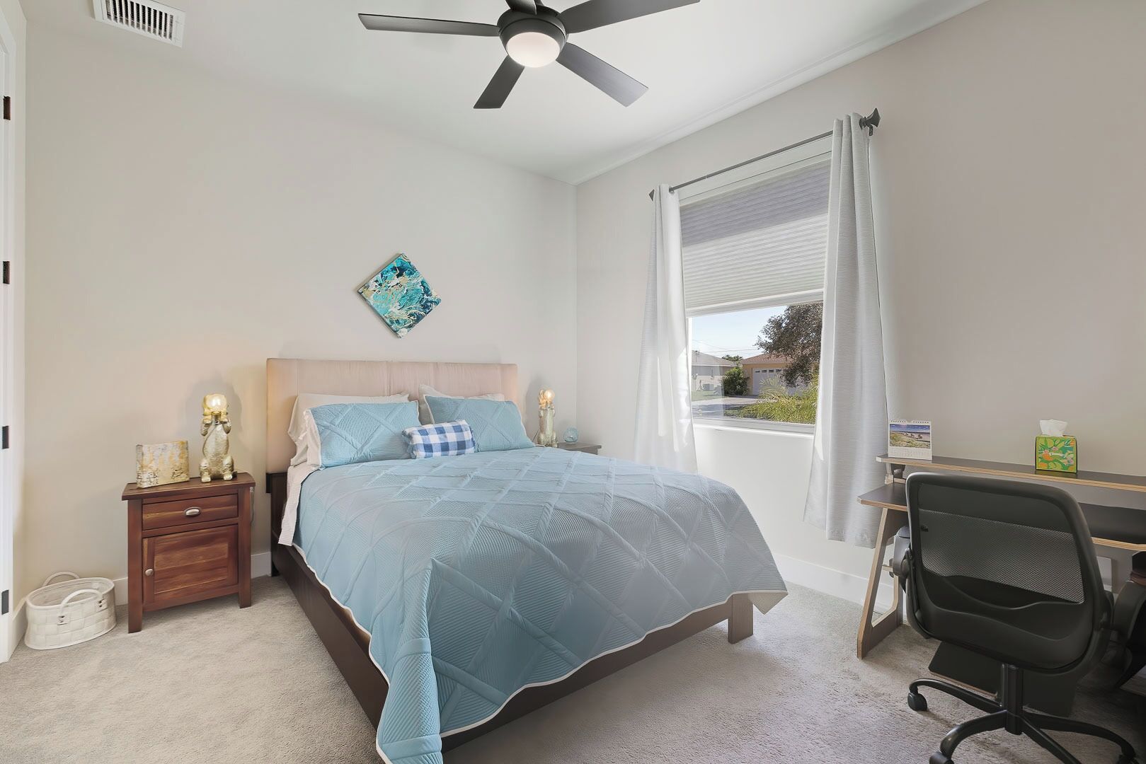 A bedroom with a bed, a desk, a chair, a ceiling fan, a window with curtains, and a wall-mounted artwork.