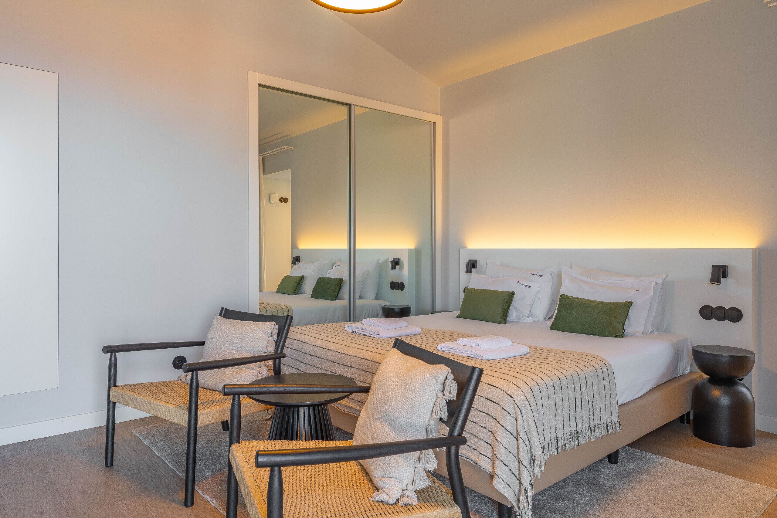 Ein modernes Hotelzimmer mit einem großen Bett, einer Sitzecke mit Sesseln, einem kleinen Tisch und einem Spiegel.