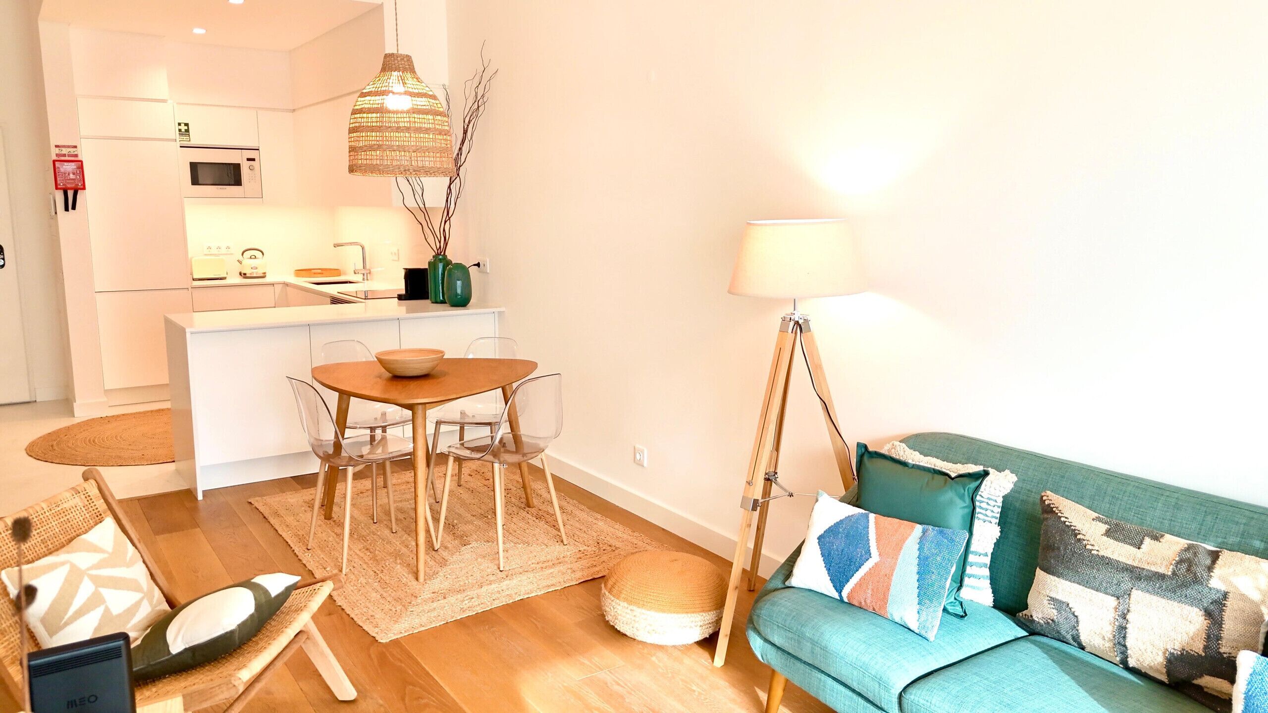 Una sala moderna con un sofá color turquesa, una mesa de comedor de madera con sillas, una zona de cocina con armarios blancos y un lámpara colgante.