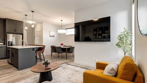 A modern kitchen with a central island, dining area with chairs, a sofa, and a TV mounted on the wall. - 7-Bedroom Getaway with AC, Hot Tub, BBQ, Garage and Pet-Friendly Amenities (Calgary)