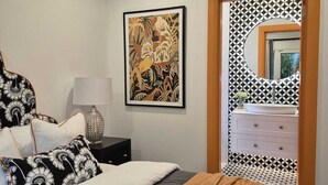 Ein Schlafzimmer mit Bett, Kissen, einem Nachttisch mit Lampe, einem gerahmten Kunstwerk und einem durch eine Tür mit ornamentalem Wandpapier einsehbarer Badezimmer.