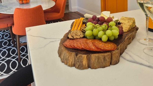 Ein Essbereich mit einem Marmortisch, orangefarbenen Stühlen, einer Holzservierplatte mit Käse, Trauben und luftgetrocknetem Fleisch. Darüber hängt eine Chandelier, und im Hintergrund ist eine Tür mit einem Gemälde zu sehen.