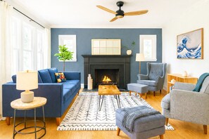 A living room with a fireplace, blue sofas, a coffee table, and a ceiling fan. - Prof's Riverside Haven Art, Hot Tub, Sleeps 10 (South Bend)