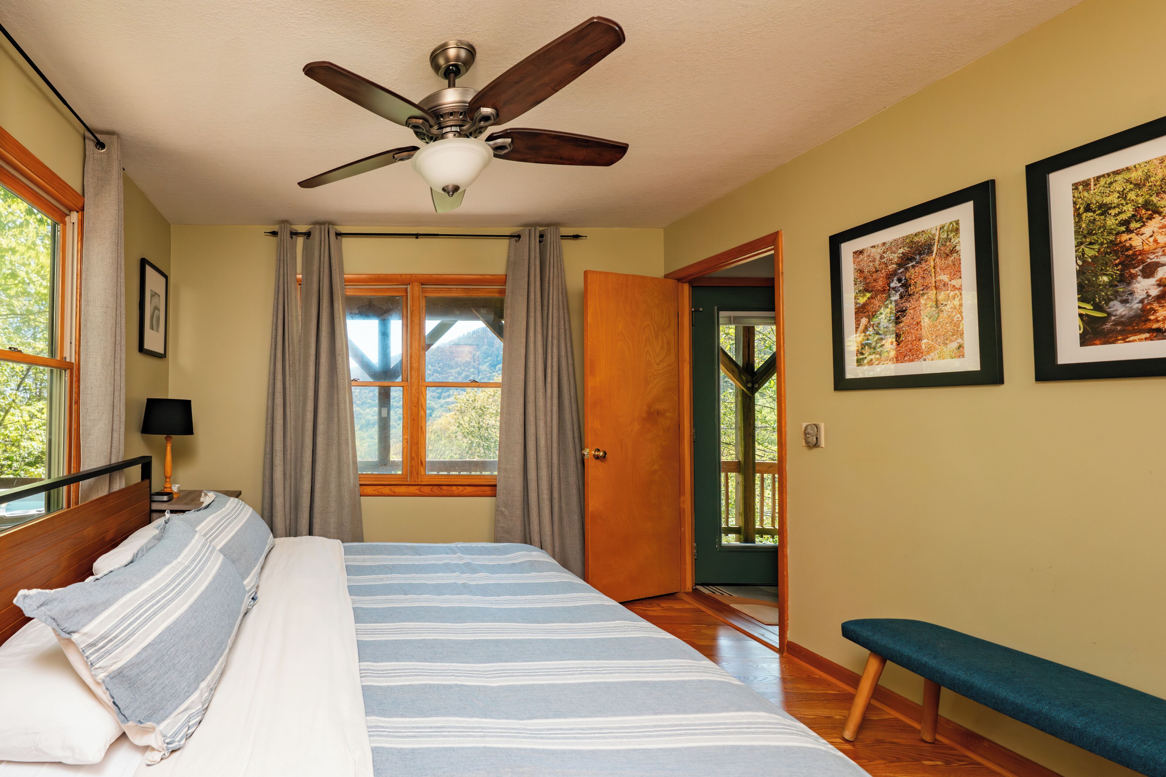 A bedroom with a bed, a ceiling fan, a window with curtains, a door, and a bench.