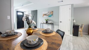 Un coin repas avec une table ronde en bois, dressée pour quatre personnes et entourée de chaises. On peut voir un vase rempli de fleurs sur la table, ainsi qu’un tableau accroché au mur.