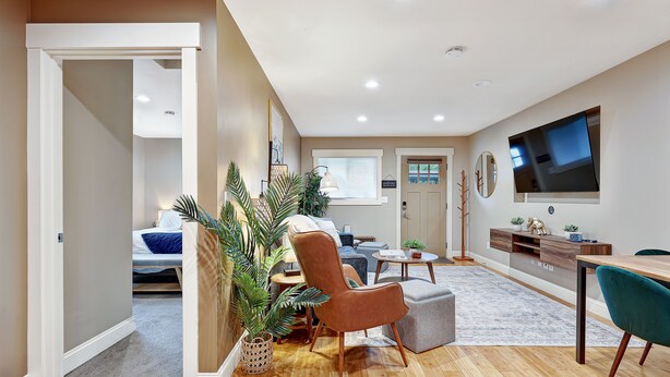 Una sala de estar moderna con un sofá, un sillón y una mesa de centro. Hay un televisor montado en la pared y una planta en maceta en la esquina.