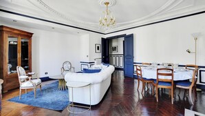 Een ruime woonkamer met een witte bank, blauwe accenten, een kroonluchter, en een eethoek met houten stoelen en een tafel.
