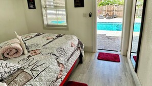 Un dormitorio con cama, ventilador de techo, ventana con persianas y vista a un patio trasero con piscina.