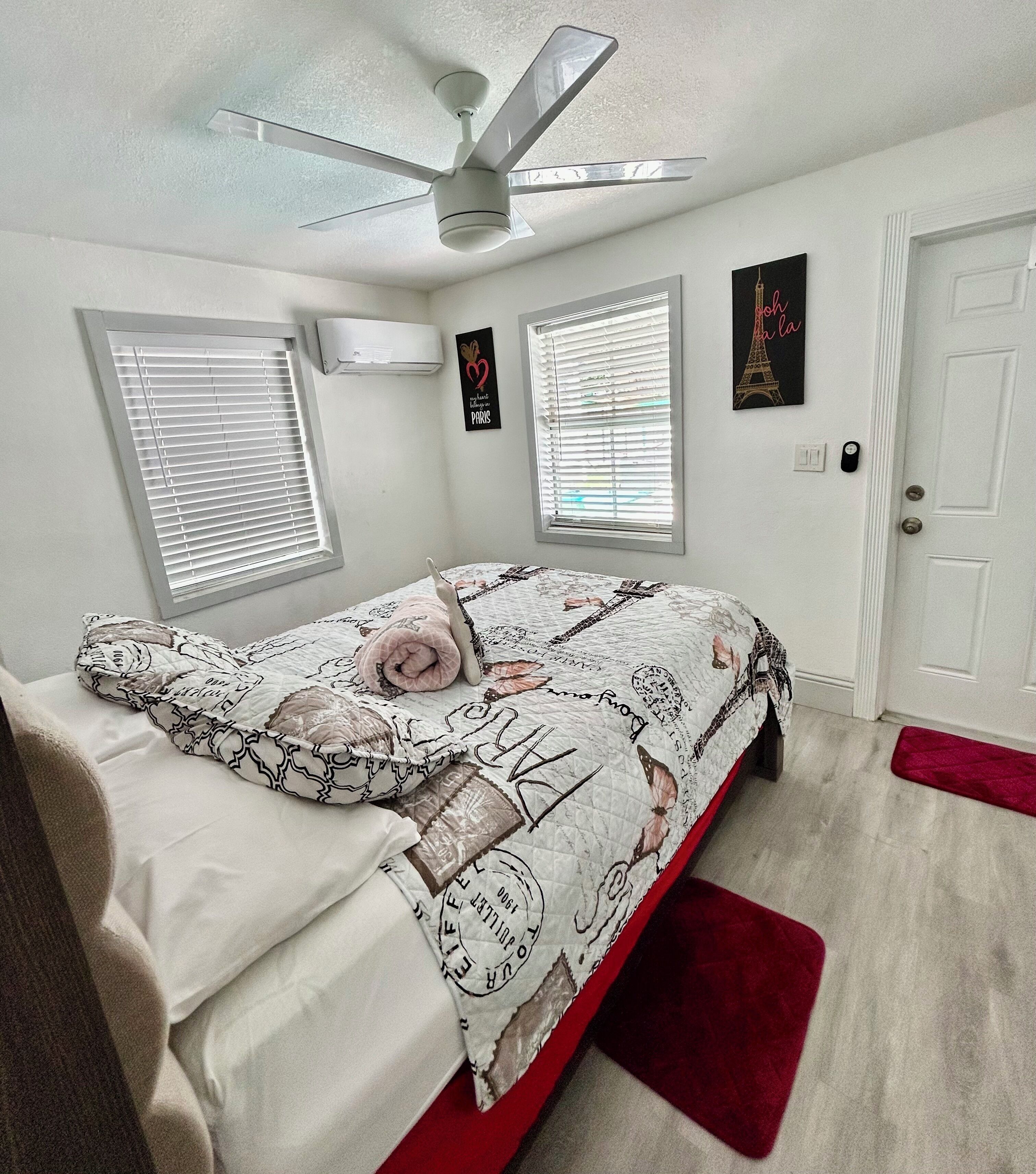A bedroom with a bed, a ceiling fan, two windows with blinds, a door, and a wall with a picture.