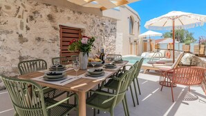 Comedor con pared de piedra, mesa de madera, sillas verdes y zona junto a la piscina con sillones y sombrillas.