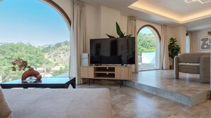 Una sala de estar moderna con un televisor de pantalla plana grande, un sofá y una mesa de centro. Se ve una ventana grande con vistas a zonas verdes y colinas.