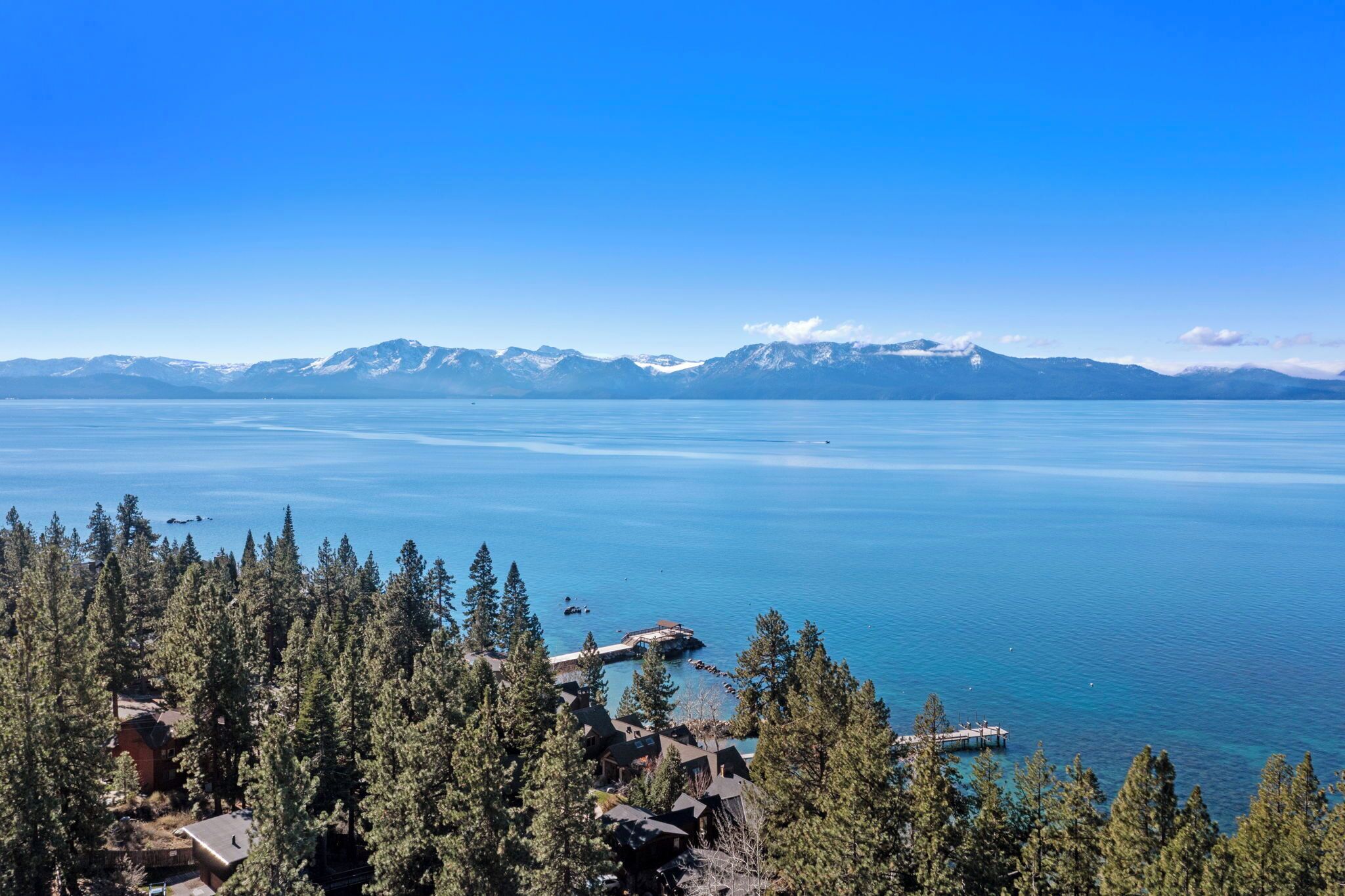 A lake with mountains in the background, surrounded by dense forest and a dock extending into the water.