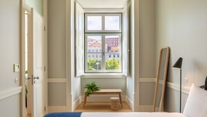 Une chambre avec une grande fenêtre, une table de chevet ornée d’une plante, une lampe de chevet et un miroir accroché au mur.