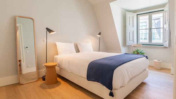 Un dormitorio con cama, mesita de noche, espejo y ventana con cortinas.