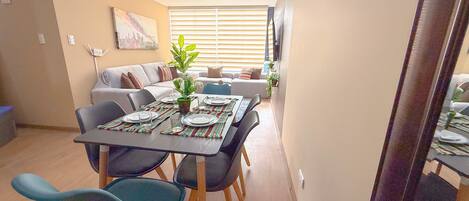 Un comedor con una mesa lista para cuatro personas, rodeada de sillas, en un apartamento moderno con un sofá y una planta al fondo.