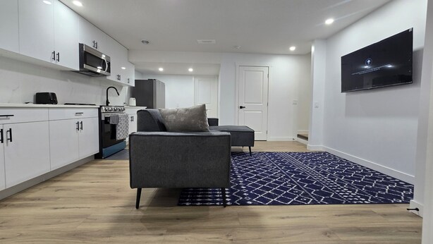 Eine moderne Küche mit weißen Schränken, einer Mikrowelle und einem Herd. Im Wohnbereich befindet sich ein graues Sofa mit einem gemusterten Teppich.