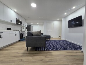 Een moderne keuken met witte kasten, een magnetron en een fornuis. In de woonkamer staat een grijze bank met een kamerbreed tapijt.