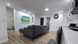 Ein modernes Wohnzimmer mit grauem Sofa, Holzboden, einem Wand-Fernseher und einer Küchenzeile mit Mikrowelle.