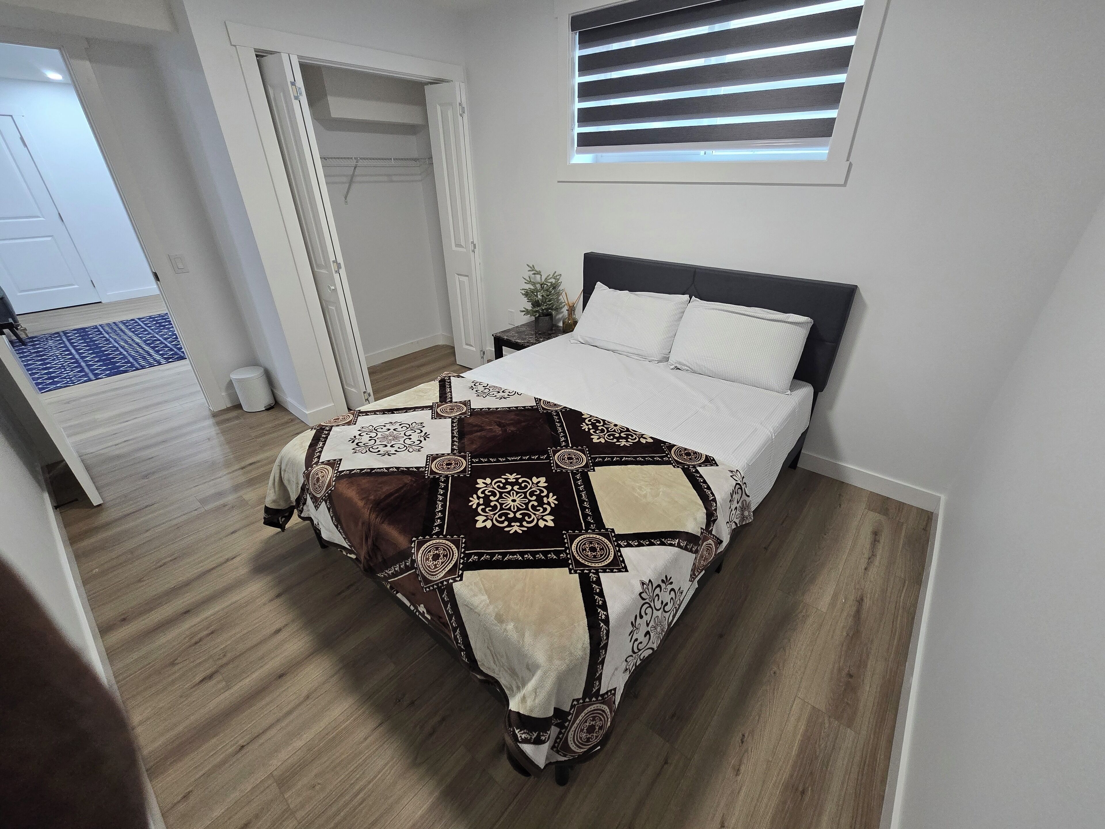 Een slaapkamer met een bed, een raam met zonwering, een klein nachtkastje en een deur naar een andere kamer.