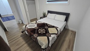 Ein Schlafzimmer mit einem Bett, einem Fenster mit Jalousien, einem kleinen Nachttisch und einer Tür zu einem anderen Raum.