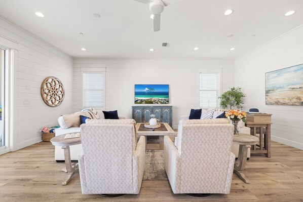 Un salon spacieux doté d’une grande fenêtre, d’un ventilateur de plafond et d’un tableau représentant une scène de plage.