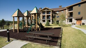 A playground with climbing structures and a picnic table, surrounded by a landscaped area and residential buildings. - WorldMark West Yellowstone condo with easy access to Yellowstone West Gate. (West Yellowstone)