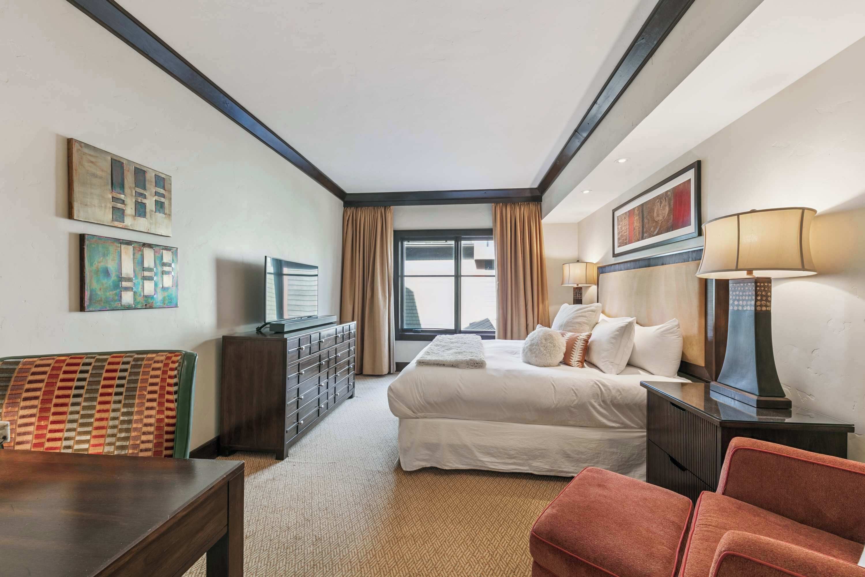 A hotel room with a bed, a desk, a chair, a dresser, and a window with curtains.