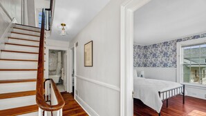 A bedroom with a wooden floor, a bed with a white bedspread, a window with a view, and a staircase with wooden railings. - Historic Mystic Maritime House - 5 min Walk to Downtown Restaurants and Seaport (Mystic)