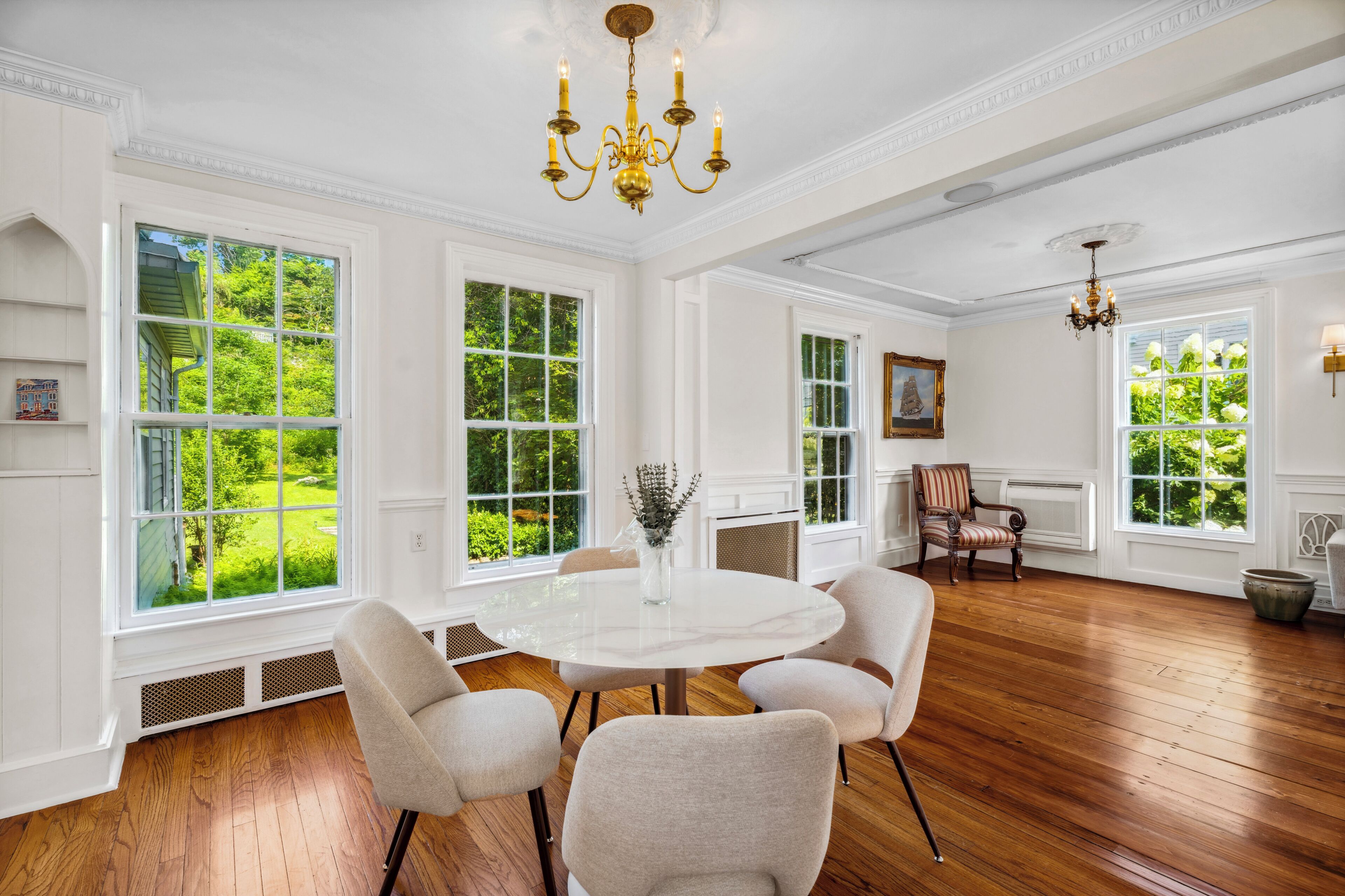 A spacious room with a round white table surrounded by four chairs, large windows with a view of greenery, and a chandelier.
