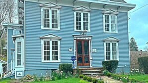 A two-story blue house with a central entrance, a small porch, and a garden with daffodils. - Historic Mystic Maritime House - 5 min Walk to Downtown Restaurants and Seaport (Mystic)