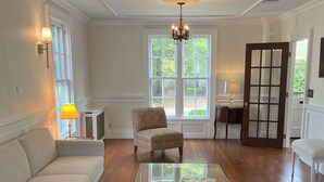 A spacious living room with hardwood flooring, a glass coffee table, a patterned armchair, a sofa, and a chandelier. - Historic Mystic Maritime House - 5 min Walk to Downtown Restaurants and Seaport (Mystic)