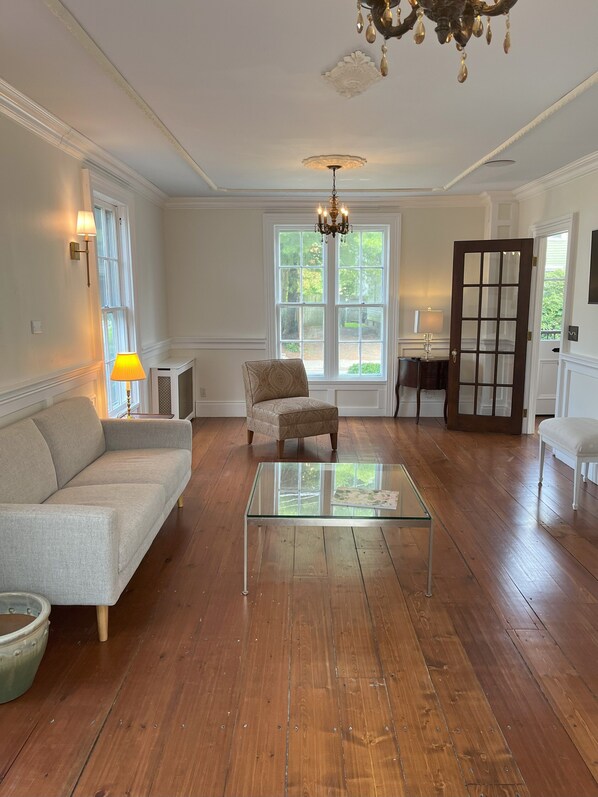 Een ruime woonkamer met houten vloeren, een glazen salontafel, een gestreept fauteuil, een bank en een kroonluchter.