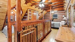 A log cabin interior with a loft bedroom, wooden beams, a ceiling fan, and a wooden table.