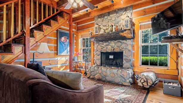 A log cabin interior with a stone fireplace, a wooden staircase, and a large window.