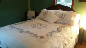Una cama con colcha y almohadas estampadas con motivos florales, una mesita de noche con lámpara y una ventana con luz diurna.
