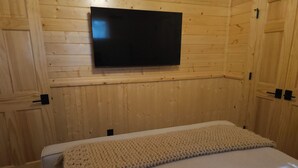 Una habitación con paredes de madera, un televisor de pantalla plana montado en la pared y una cama con colchón blanco y manta de punto color beige.