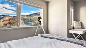 Un dormitorio con un ventanal grande que ofrece vistas a la montaña, un telescopio sobre un trípode y una pequeña mesa blanca con un cojín.