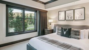 Ein Schlafzimmer mit einem großen Fenster, einem Bett mit kariertem Überwurf, einem Nachttisch mit Lampe und gerahmten Bildern an der Wand.