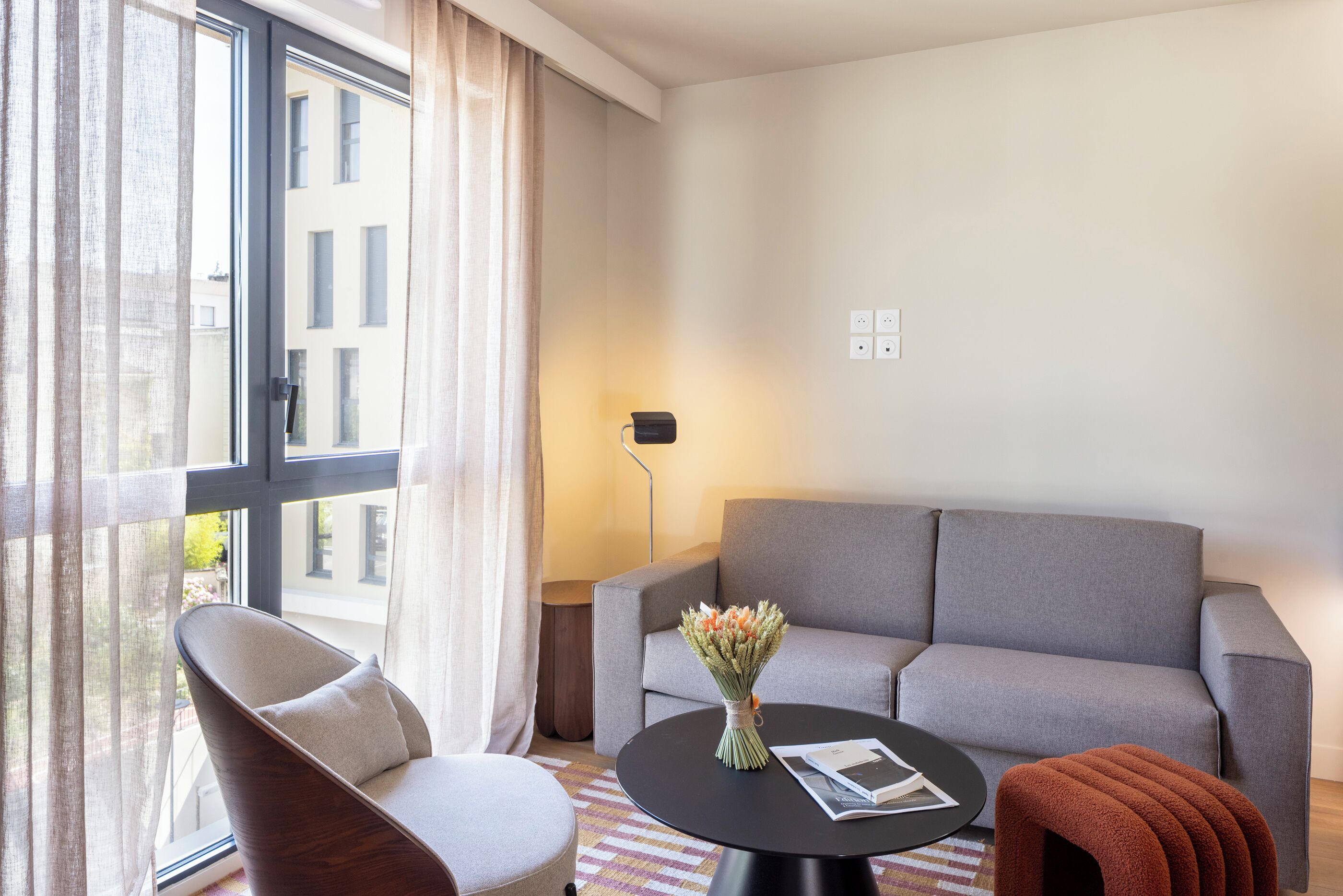 Une chambre d’hôtel moderne avec un canapé gris, une table ronde noire, une chaise et une fenêtre avec des rideaux.