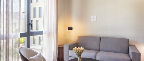A modern hotel room with a grey sofa, a round black table, a chair, and a window with curtains.