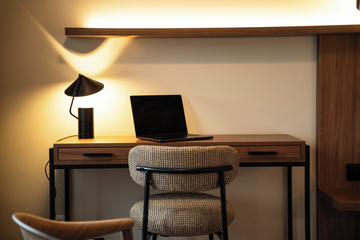 Ein moderner Arbeitsplatz mit einem Laptop auf einem Holztisch, einer Lampe und einem gemusterten Stuhl.