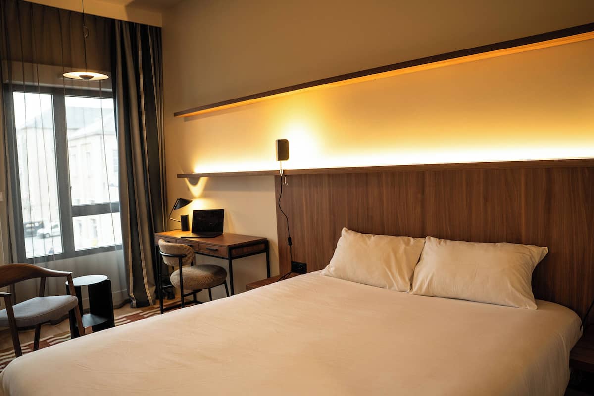 Une chambre d’hôtel avec un grand lit, un bureau avec une chaise, une lampe et une vue sur la ville par la fenêtre.