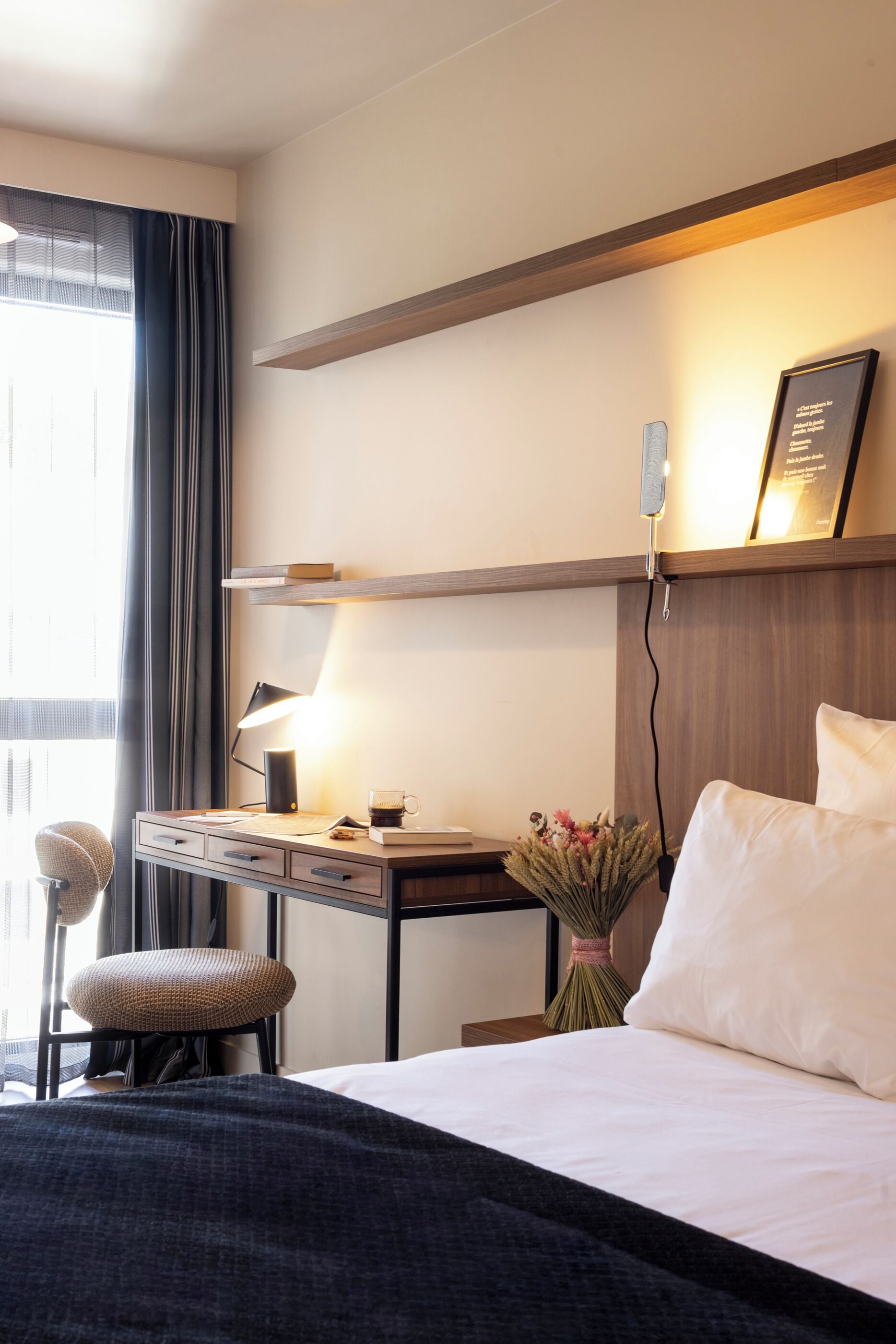A hotel room with a bed, a desk with a lamp, a chair, and a vase with flowers.