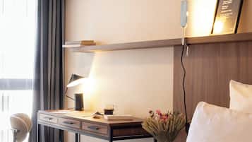 A hotel room with a bed, a desk with a lamp, a chair, and a vase with flowers.